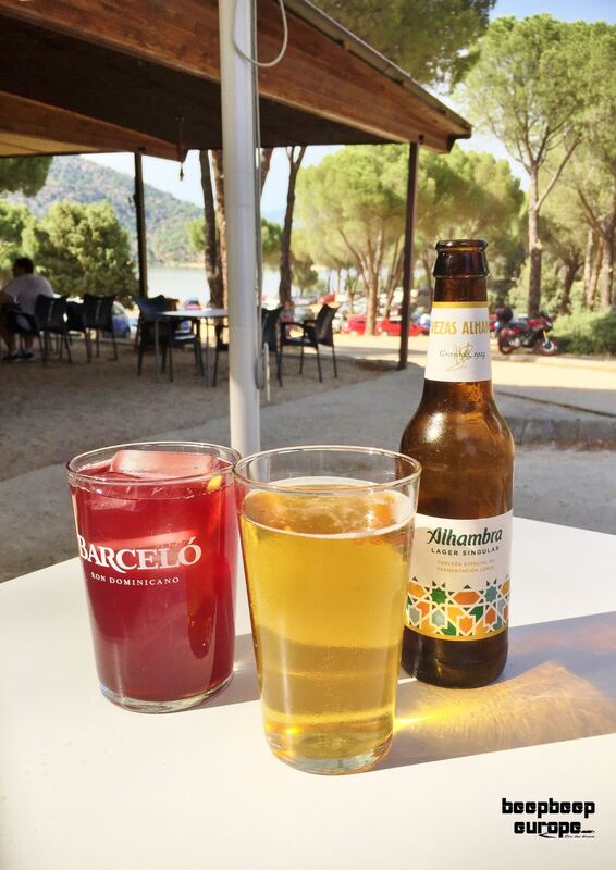 Two glasses, a beer bottle on a table. One filled with beer, one with a red drink in an outdoor cafe with a lake in the background.