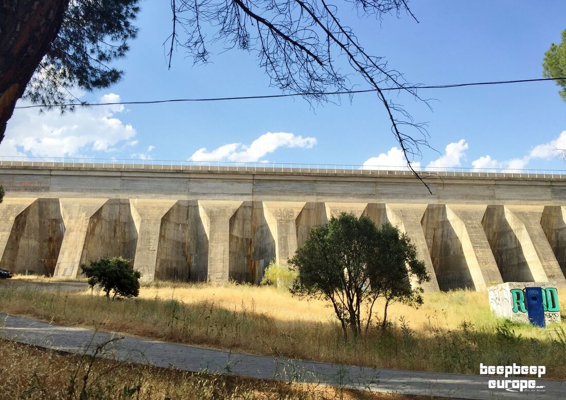 In the countryside at the bottom of a cement dam wall with many arches, surrounded by trees and dry shrubs and vegetation.