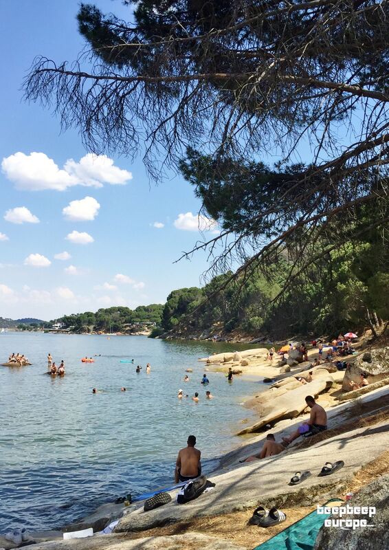 A number of people are dotted along the shoreline of a lake, sitting, sunbathing on granite slabs or enjoying a dip in the water.