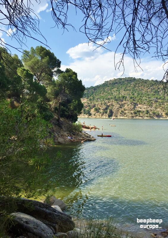 Deep green rocky hills filled with trees surround a stunning lake. Several people are enjoying a dip, sailing and kayaking.