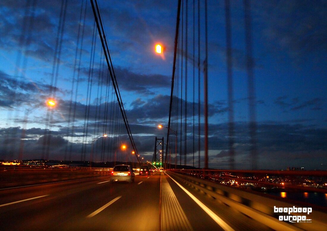 Driving across a suspension bridge at night, with cars ahead and city lights below and dark blue, cloudy skies above.