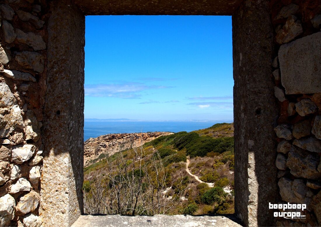 Looking through a stone-walled window onto a green clifftop. The blue ocean is on the horizon with vibrant blue skies above.Picture