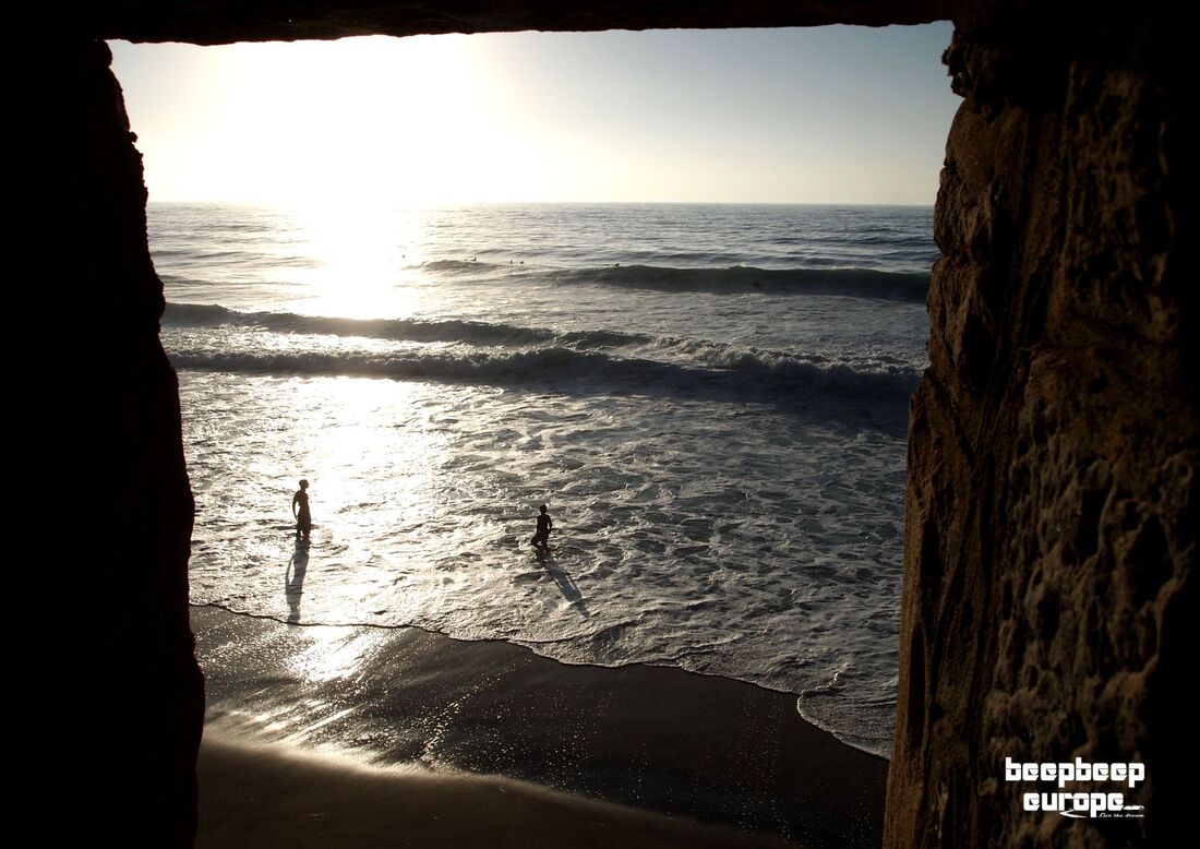 Looking through a rock-made window onto a dark, shadowy beach scene of two people in the water with waves rolling in.