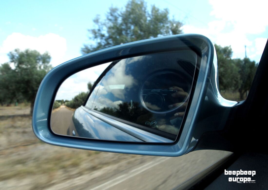 On the road in the countryside looking into left wing mirror with reflection of the side of a light blue car and road behind.