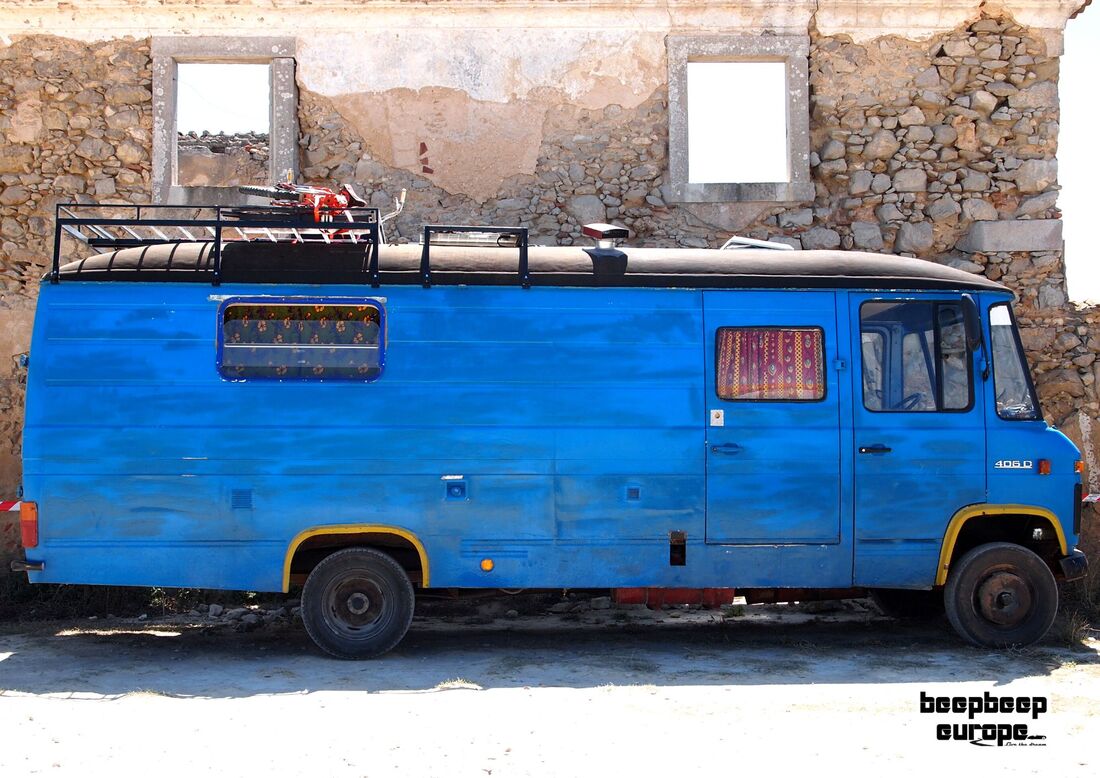 Side on of retro blue long wheelbase motorhome with coloured curtains parked in front of a stone-walled derelict building.