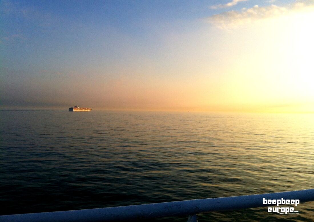 View from the deck onboard a ship in the ocean. A container ship is sailing away on the horizon whilst the sun is setting.