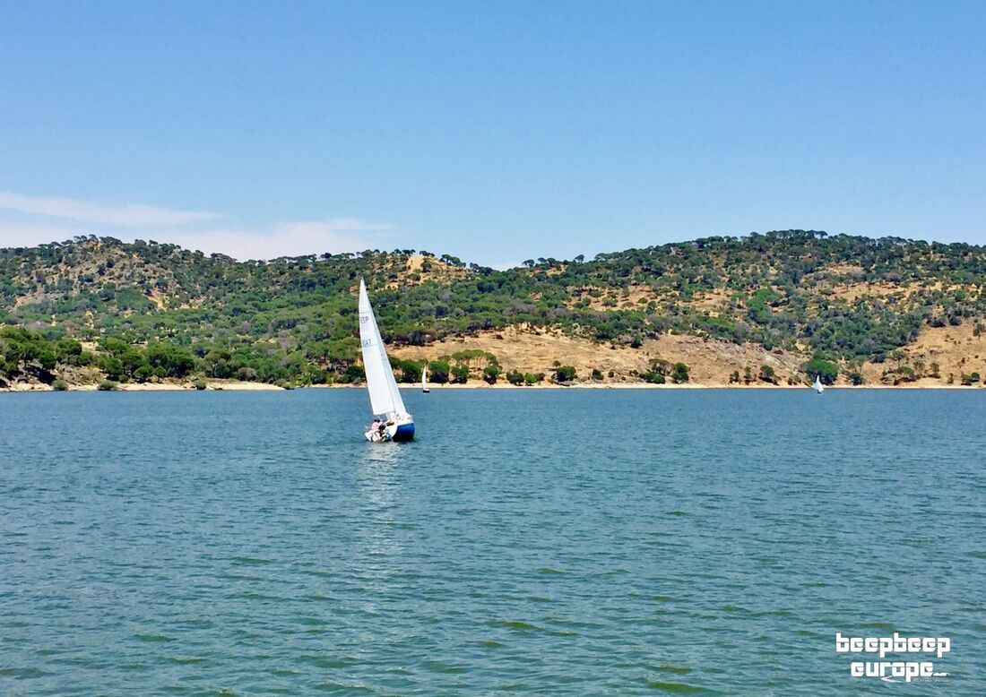 A small yacht is sailing around a lake on beautiful blue waters, surrounded by several very green, rocky hills in the background.